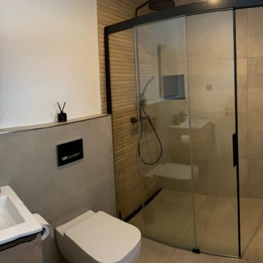 Modernes Badezimmer mit bodengleicher Dusche, schwarzer Glasabtrennung, WC und Waschtisch in Holzoptik.