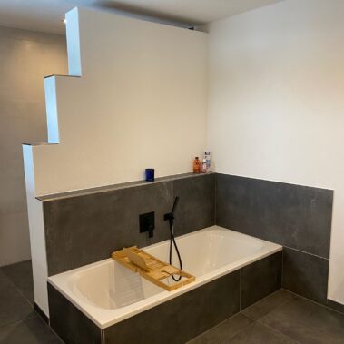Großzügiges Badezimmer mit eingelassener Badewanne, dunklen Fliesen und moderner Raumtrennung.