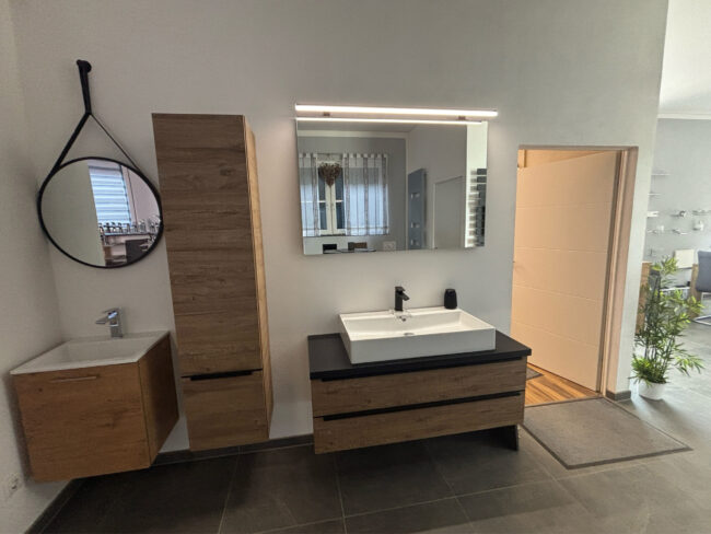 Ausstellung eines modernen Badezimmer-Waschplatzes mit Holzoptik, beleuchtetem Spiegel, Hochschrank und schwarzer Arbeitsplatte.