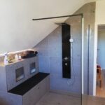 Bodengleiche Dusche im Dachgeschoss mit großformatigen Betonoptik-Fliesen, schwarzer Regendusche, Glasabtrennung und integrierter Sitzbank mit Ablagenischen.