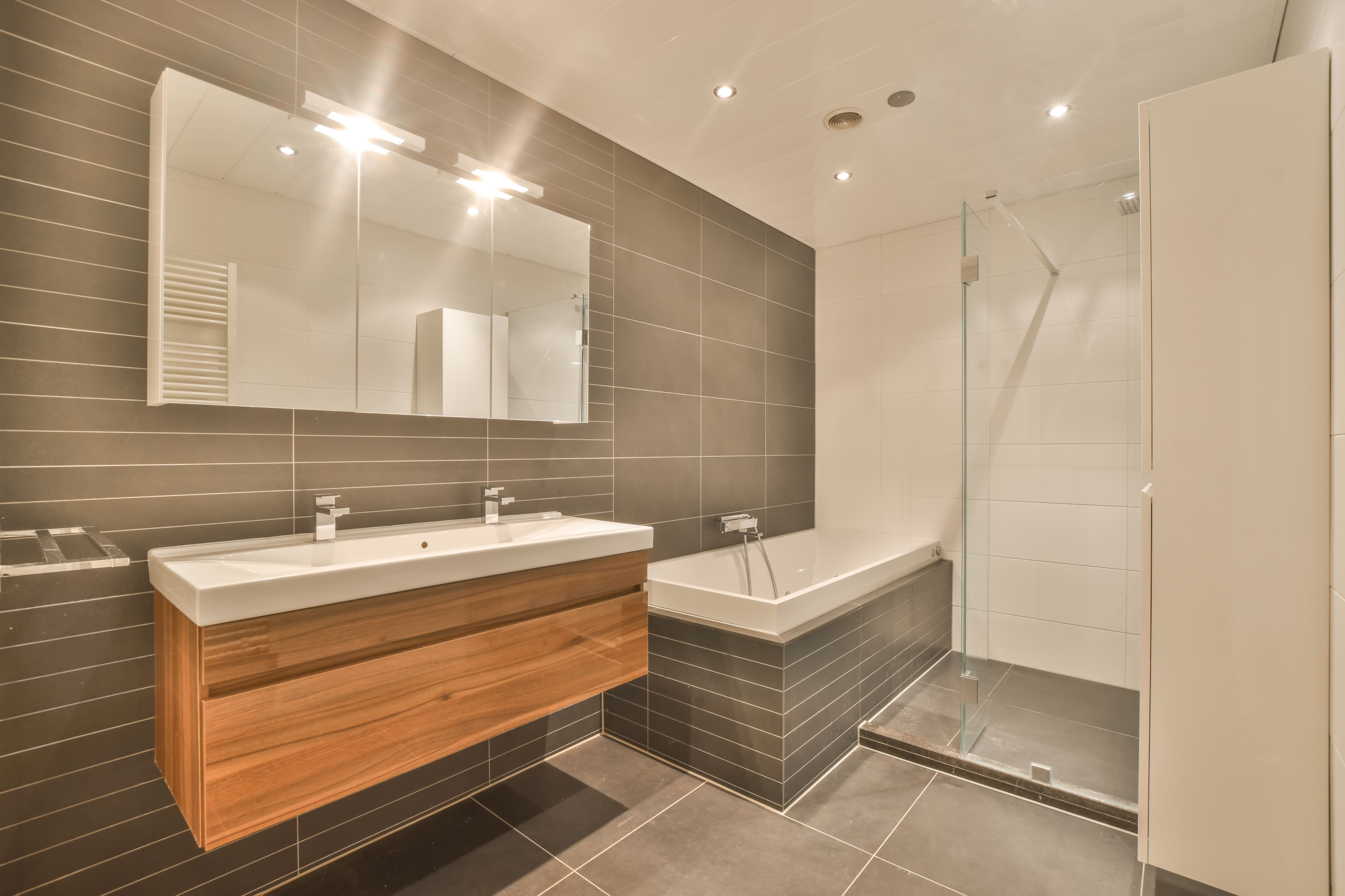 Zeitloses Badezimmer mit Doppelwaschtisch in Holzoptik, integrierter Badewanne, bodengleicher Glasdusche und dunklen Wandfliesen.